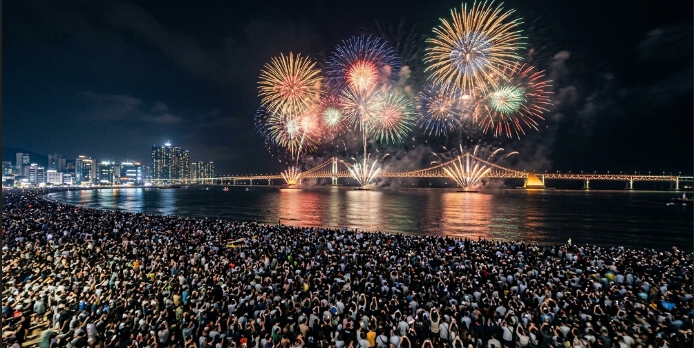 광안대교 위로 화려한 대형 불꽃놀이가 터지고 있는 밤하늘 아래, 광안리 백사장을 발 디딜 틈 없이 빽빽하게 가득 채운 수많은 인파의 모습. 낭만적인 밤하늘과 대비되는 극심한 혼잡도를 보여준다.